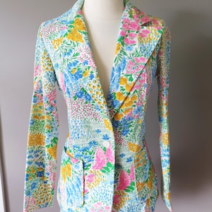 May include: A floral blazer with a colorful pattern of pink, yellow, blue, and green flowers on a white background. The blazer has a single-button closure, notched lapels, and two front pockets. The sleeves are long.
