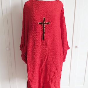 Op de afbeelding: Een rode, getextureerde tuniek met lange mouwen en een boothals. Een bruin kruis is gecentreerd op de voorkant. Het kledingstuk wordt gepresenteerd op een mannequin tegen een witte achtergrond.