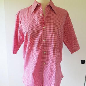 Può includere: Una camicia a maniche corte con bottoni e un motivo a quadretti vichy rosso e bianco. La camicia presenta un colletto a punta, un'abbottonatura frontale con cinque bottoni bianchi visibili e un orlo dritto, esposta su un busto di manichino.