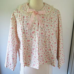 May include: A quilted jacket featuring a cream base with a small-scale floral pattern in pink and blue. The jacket has long sleeves, a rounded collar with lace trim, and a light pink ribbon tied in a bow at the center of the neckline.