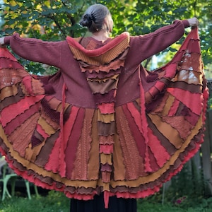 May include: A patchwork cardigan in red, brown, and orange hues. The hooded, open-front sweater has a distinctive design with layered fabric panels and a ruffled trim. The textured garment is displayed outdoors, showcasing its artistic style. The cardigan is a unique piece.
