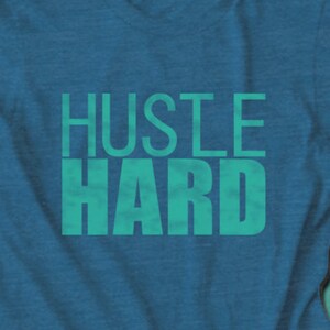 May include: A teal blue t-shirt with the text "Hustle Hard" printed in a teal green color.