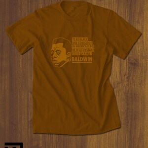 May include: A brown t-shirt with a black and white illustration of James Baldwin. The text on the shirt reads "To be black and conscious in America is to be in a constant state of rage! Baldwin."