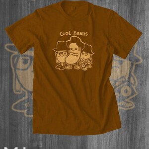 May include: A brown t-shirt with a graphic of three cartoon beans wearing hats and sunglasses. The text "Cool Beans" is printed above the beans.