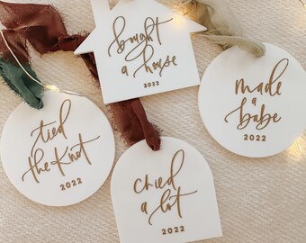 May include: Four white ceramic ornaments with gold lettering. The ornaments are shaped like a house, a circle, an arch, and a circle. The text on the ornaments reads "bought a house 2022", "tied the knot 2022", "cried a lot 2022", and "made babe 2022".