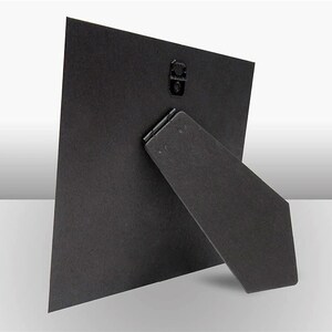 May include: Black cardboard photo frame with a built-in easel stand on the back. The frame is designed to stand upright on a table or shelf.