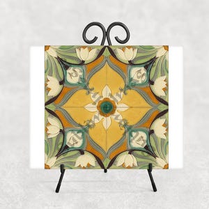 May include: A decorative tile with a floral design in yellow, green, and white, displayed on a black metal easel. The tile features a symmetrical pattern with stylized flowers and leaves, creating a vintage aesthetic.