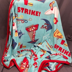 May include: A turquoise fleece blanket with a red crocheted border. The blanket features bowling-themed graphics, including bowling pins, bowling balls, and the word "STRIKE!" in red. Other graphics include a cafe sign, a hamburger, and a soda.