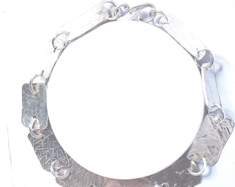 May include: A silver-toned bracelet with a geometric design. The bracelet features rectangular links connected by small, circular links. The metal has a textured appearance, and the bracelet is displayed on a white surface.