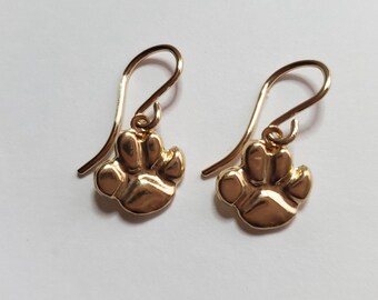 May include: A pair of gold-colored drop earrings. Each earring features a paw print charm suspended from a curved ear wire. The earrings are displayed against a white background.