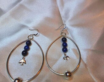 May include: A pair of silver-toned teardrop-shaped earrings featuring a central vertical strand of three dark blue spherical beads and a small silver bird charm. A single silver-toned bead is attached to the bottom of each hoop.
