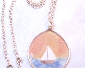 May include: A silver chain necklace with a round pendant featuring a watercolor painting of a sailboat on the water with a sunset sky.