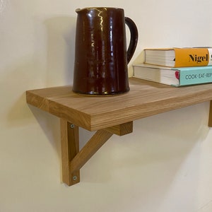May include: A wooden shelf with a brown ceramic jug and two cookbooks on top. The shelf is mounted to the wall with two wooden brackets.