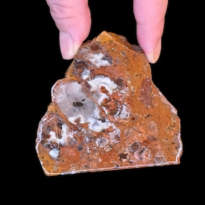 May include: A rough-cut, natural stone with a mix of orange, brown, and white patterns. The stone has a unique shape and texture, with visible variations in color and veining. The stone is held between two fingers against a black background.