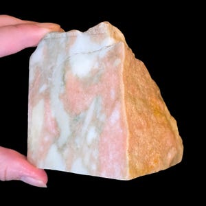 May include: A rough-cut mineral specimen featuring a marbled pattern of soft pink, creamy white, and muted grey tones. The stone has a jagged, uneven top edge and a solid, blocky shape with a smooth, polished front face displaying natural veining.