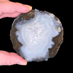 May include: A circular agate slice featuring a translucent, pale blue-grey center with a crystalline, textured core. The stone is surrounded by a dark, earthy brown outer rim with a rough, natural edge, showcasing distinct concentric banding.