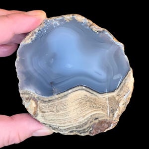 May include: A circular agate slice featuring a smooth, translucent light blue center with subtle banding, transitioning into a textured, earthy tan and brown outer rim with distinct mineral layers and a rough, natural edge against a black background.