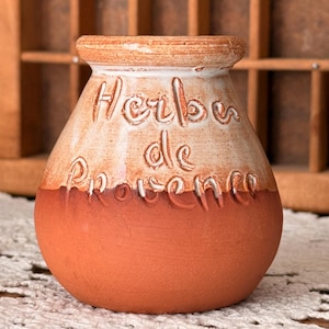 May include: A small, round, terracotta-colored ceramic jar with a wide opening. The upper portion features a white glazed finish with the embossed text "Herbes de Provence" in a script font, while the lower portion remains unglazed terracotta.