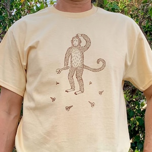 May include: Cream-colored t-shirt featuring a brown illustration of a monkey holding a wrench, scratching its head, and surrounded by screws. The design is centered on the shirt, with the monkey's tail curled.