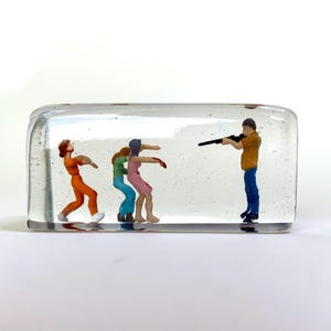 May include: A clear glass block with a scene inside. Three figures are walking towards a man holding a rifle. The figures are painted in orange, blue, and pink. The man is wearing a yellow shirt and blue jeans.