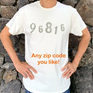 May include: A white short-sleeved t-shirt featuring the grey printed numbers 96816 across the chest. Below the numbers, the text Any zip code you like! is printed in orange. The shirt has a crew neckline and a standard fit for casual wear.
