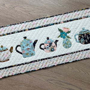 Peut inclure: Un chemin de table matelassé blanc avec une bordure noire et dorée. Le chemin de table présente cinq théières appliquées et un vase de fleurs dans une variété de motifs floraux.