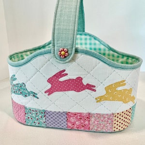May include: A quilted Easter basket with a green handle and a pink, yellow, and blue patchwork design. The basket features three bunny silhouettes in pink, yellow, and blue.