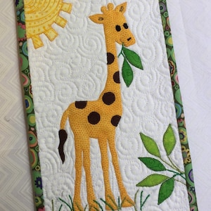 May include: A quilted wall hanging featuring a giraffe with brown spots, eating green leaves. The giraffe is set against a white background with a yellow sun and green grass. The border is a colorful floral pattern.