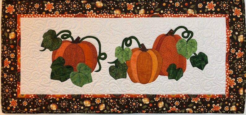 K&ouml;nnte beinhalten: Ein wei&szlig;er gesteppter Tischl&auml;ufer mit braunem Blumenrand und drei orangefarbenen K&uuml;rbissen mit gr&uuml;nen Bl&auml;ttern und Ranken.