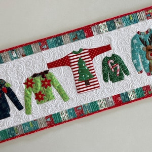 Może przedstawiać: Pikowany bieżnik stołowy z białym tłem i czerwoną obwódką. Pięć różnych świątecznych swetrów jest naszytych na bieżniku, każdy z unikalnym wzorem. Swetry są zielone, czerwone i niebieskie z różnymi motywami świątecznymi.