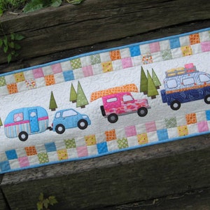 May include: A white quilted table runner with a blue border features a row of colourful vehicles, including a camper, a car, a jeep with a canoe on top, and a van. The vehicles are surrounded by green trees and a patchwork of colourful squares.