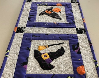 May include: A purple and white quilted table runner with a Halloween theme. The runner features four squares with witch hats, brooms, and pumpkins. The squares are outlined in white and black fabric.