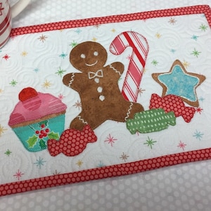 Peut inclure: Un set de table matelassé sur le thème des fêtes. Il présente un bonhomme en pain d'épices, une canne en bonbon, une étoile, un cupcake et des bonbons appliqués sur un fond blanc avec une bordure à pois rouges. Le fond a de petits accents d'étoiles et de flocons de neige.