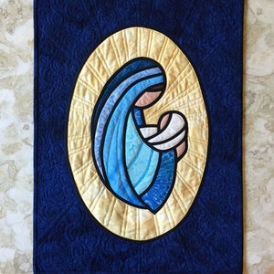 Könnte beinhalten: Ein blauer und gelber gesteppter Wandbehang, der die Jungfrau Maria darstellt, die das Jesuskind hält. Das Bild ist in einem schwarzen Oval gerahmt.