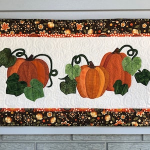 K&ouml;nnte beinhalten: Ein wei&szlig;er gesteppter Wandbehang mit braunem Blumenrand. In der Mitte befinden sich drei orangefarbene K&uuml;rbisse mit gr&uuml;nen Bl&auml;ttern und Ranken.