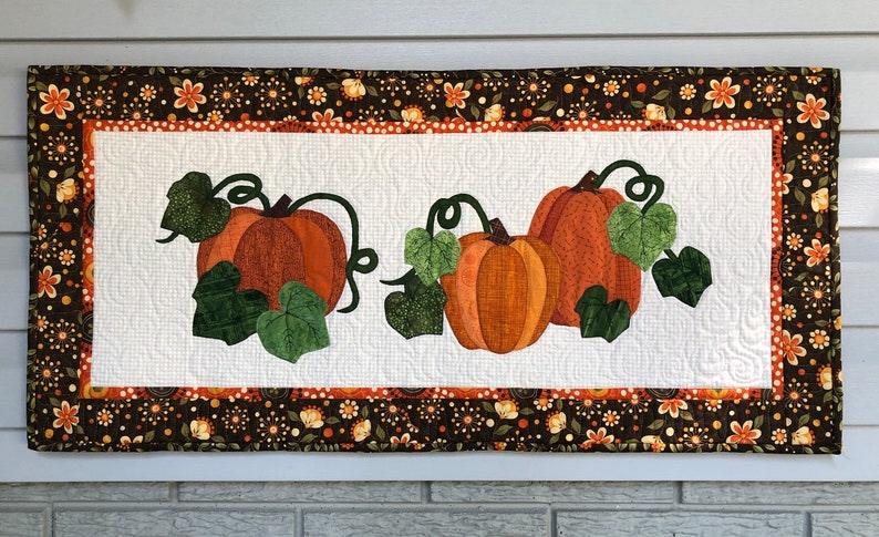 K&ouml;nnte beinhalten: Ein wei&szlig;er gesteppter Wandbehang mit braunem Blumenrand. In der Mitte befinden sich drei orangefarbene K&uuml;rbisse mit gr&uuml;nen Bl&auml;ttern und Ranken.