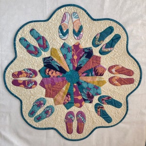 May include: A white fabric wall hanging with a circular design in the centre. The design is made up of various fabric scraps in shades of purple, pink, blue, and green. The fabric scraps are arranged in a radial pattern, creating a flower-like design. Around the centre design are 10 flip-flop shapes made of fabric in shades of blue and green.