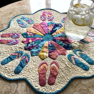 Puede incluir: Un camino de mesa acolchado blanco y azul con un diseño floral en el centro. El camino de mesa está decorado con formas de chanclas en varios colores, incluyendo rosa, azul y amarillo.