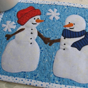 May include: A blue quilted coaster with a white border and white polka dots features two snowmen holding hands. The snowman on the left has a red hat and the snowman on the right has a blue and white striped scarf.