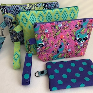 Pode incluir: Quatro bolsas de zíper de tecido diferentes com vários padrões. As bolsas são em tons de verde, azul, rosa e roxo. Os padrões incluem paisley, diamantes, guaxinins e bolinhas.