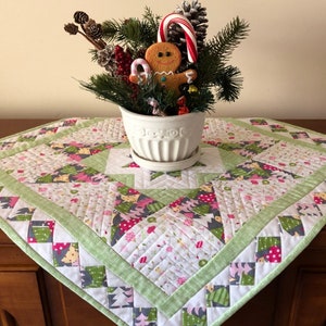 Pode incluir: Um corredor de mesa acolchoado verde e branco com um padrão de árvore de Natal. O corredor de mesa é decorado com um vaso de cerâmica branco com um homem de gengibre e bengalas de doces.