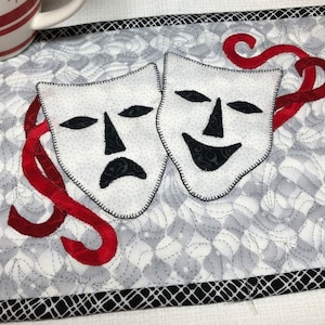 May include: A white quilted coaster with a black and white patterned border. The coaster features two fabric masks, one smiling and one frowning, with red fabric ribbons.