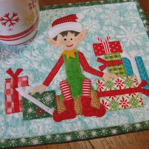 May include: A green and red Christmas elf with a white hat and a white beard is holding a stack of wrapped presents. The elf is sitting on a blue and white background with snowflakes.