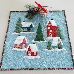 Op de afbeelding: Een gewatteerde kersttafeltopper met geappliqueerde huizen en groenblijvende bomen op een lichtblauwe achtergrond. De huizen hebben witte, met sneeuw bedekte daken en zijn in tinten rood, blauw en blauwgroen. Een decoratieve rand en een geruite ornament maken het ontwerp compleet.