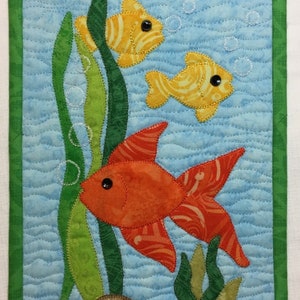 May include: A quilted wall hanging featuring three fish swimming in a blue ocean scene. The fish are orange, yellow, and red with black eyes. The background includes green seaweed and brown rocks.