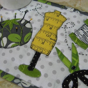 Puede incluir: Una alfombrilla de costura de tela blanca con un alfiletero gris y verde, una cinta métrica amarilla, unas tijeras verdes y negras y una bobina de hilo verde y blanca. La alfombrilla tiene un fondo blanco con lunares grises.