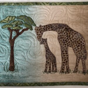 May include: A quilted wall hanging featuring a giraffe family and a tree. The giraffes have brown and tan patterned spots. The background is a mix of blue and tan, with a green border. The tree has a brown trunk and green leaves.