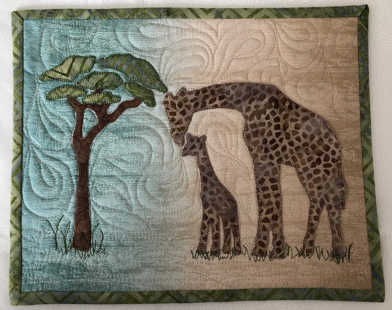 May include: A quilted wall hanging featuring a giraffe family and a tree. The giraffes have brown and tan patterned spots. The background is a mix of blue and tan, with a green border. The tree has a brown trunk and green leaves.