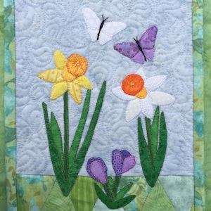 Könnte beinhalten: Ein gesteppter Wandbehang mit einer Frühlingsblumen-Szene mit gelben und weißen Narzissen, violetten Krokusse und zwei Schmetterlingen, einem weißen und einem violetten, auf hellblauem Hintergrund.