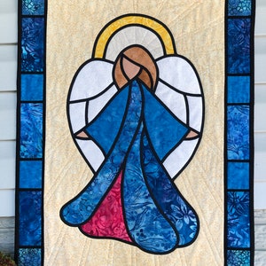 May include: A stained glass style quilted wall hanging featuring an angel with blue and red robes and white wings. The angel has a halo and is surrounded by a yellow and white background.